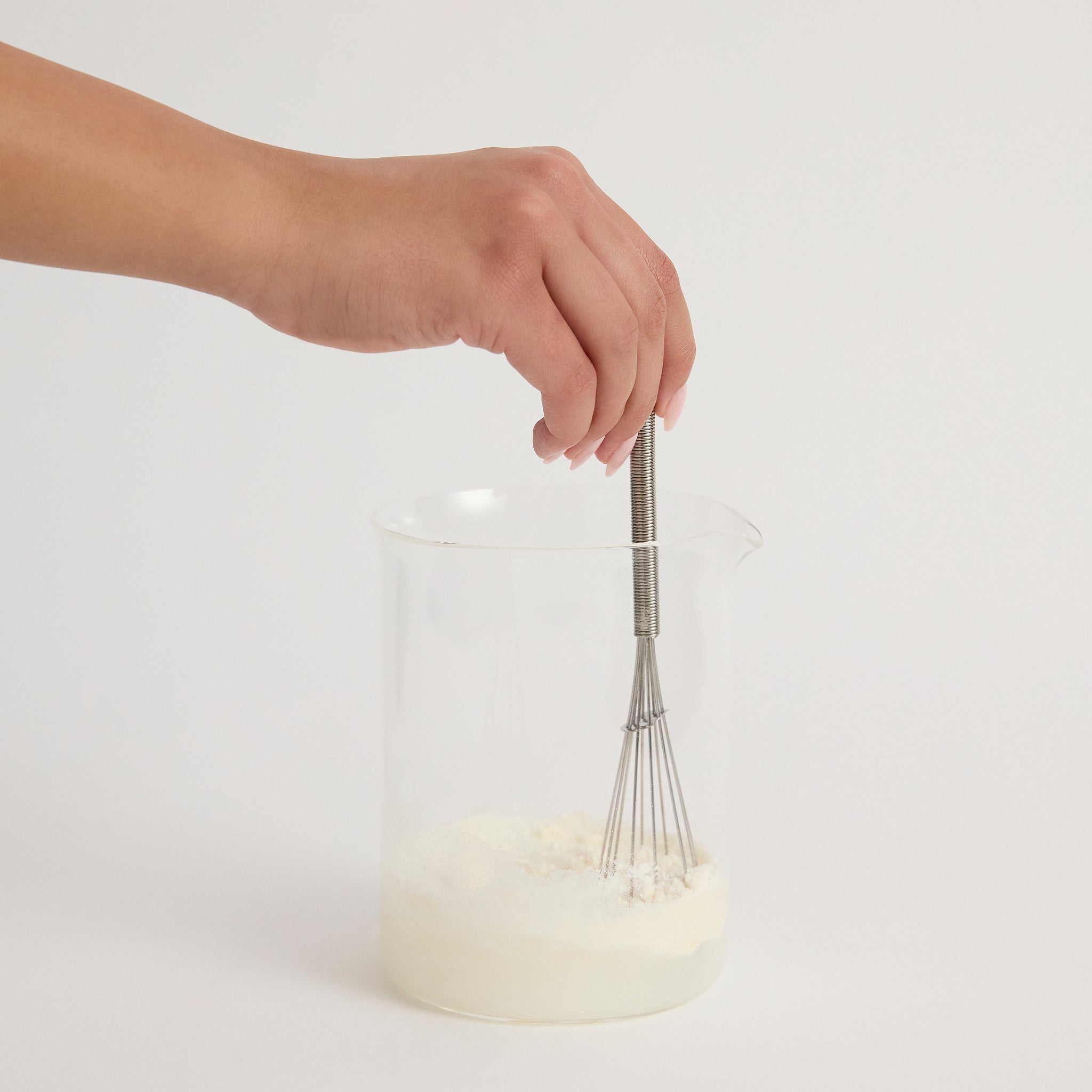 Hand using a whisk to stir a Nutritional Milk in a clear container on a light grey background